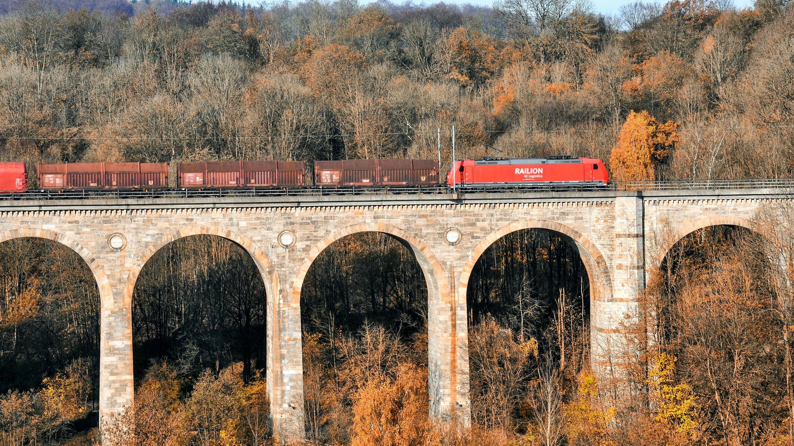 Red cargo train crossing Altenbeken Viaduct amidst autumn foliage.